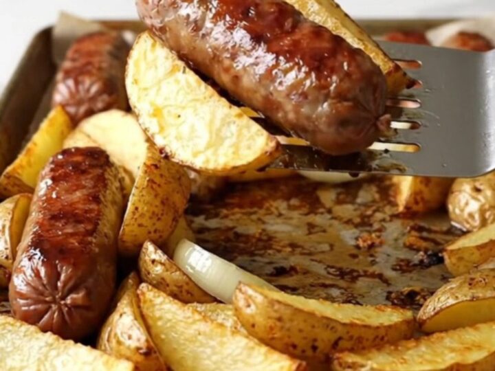 A sheet pan filled with golden-brown bratwurst, crispy potato wedges, and soft onions.