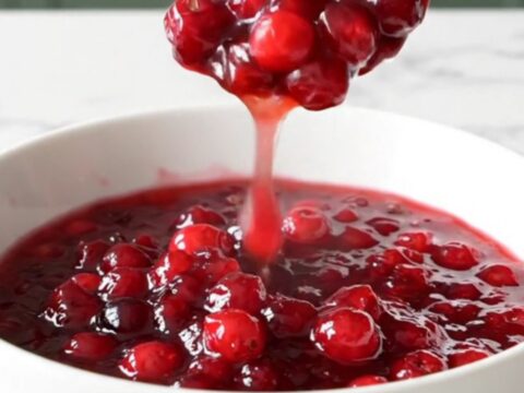 A shallow white bowl filled with chunky, deep ruby-red cranberry apple sauce, with a spoon lifting a portion.