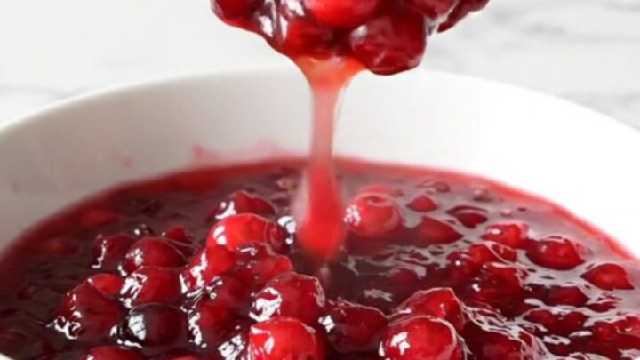 A shallow white bowl filled with chunky, deep ruby-red cranberry apple sauce, with a spoon lifting a portion.