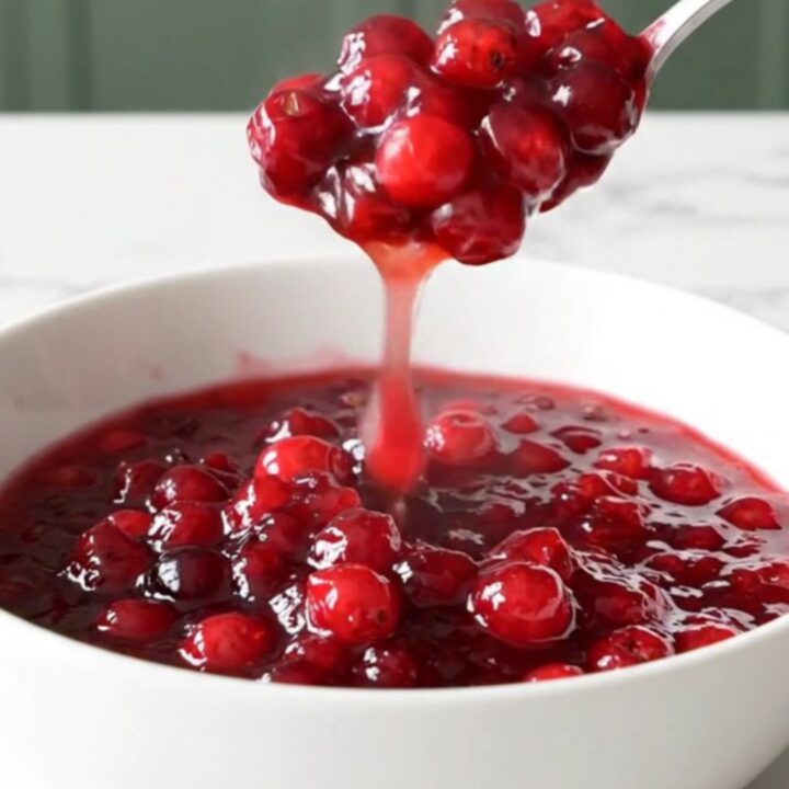 A shallow white bowl filled with chunky, deep ruby-red cranberry apple sauce, with a spoon lifting a portion.