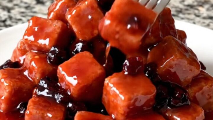 Slow cooker filled with glossy, red grape jelly glazed ham cubes on a speckled granite countertop.