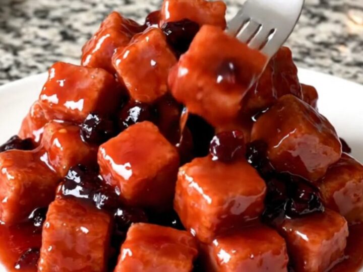 Slow cooker filled with glossy, red grape jelly glazed ham cubes on a speckled granite countertop.