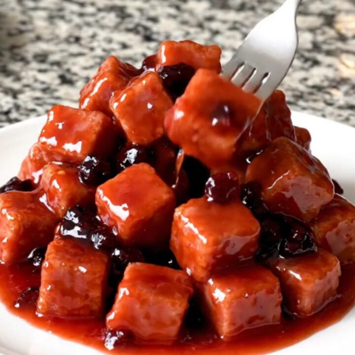 Slow cooker filled with glossy, red grape jelly glazed ham cubes on a speckled granite countertop.