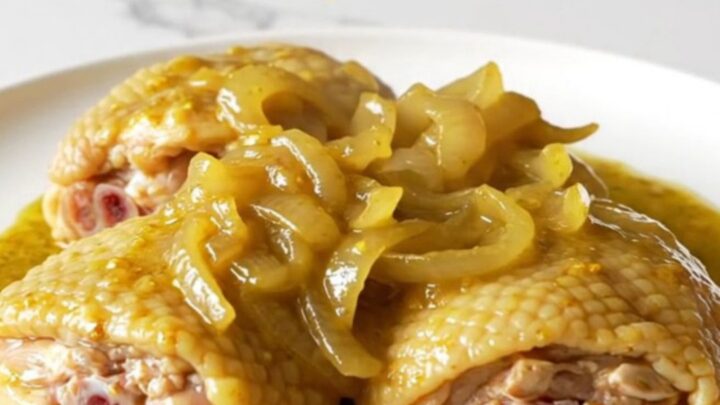 A close-up of tender, slow-cooked bone-in chicken thighs covered in a rich, golden mustard sauce with soft onions.