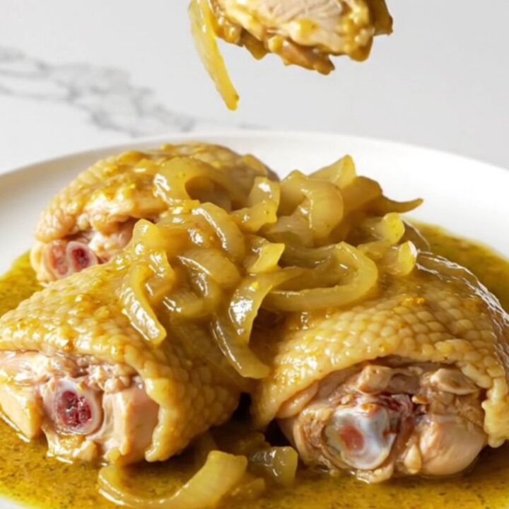A close-up of tender, slow-cooked bone-in chicken thighs covered in a rich, golden mustard sauce with soft onions.
