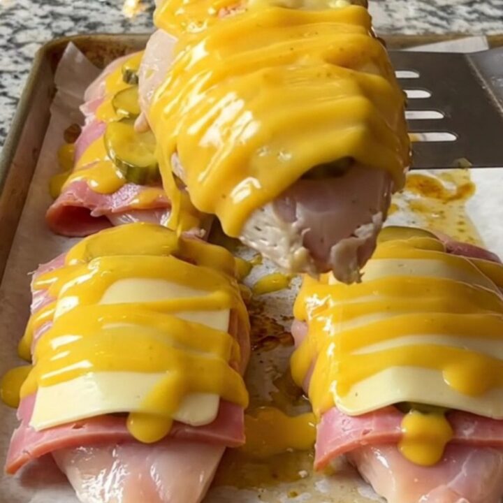 A sheet pan filled with golden-baked chicken breasts topped with ham, pickles, and melted Swiss cheese.