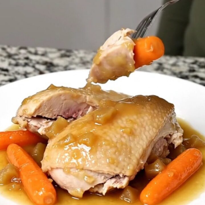 A close-up of tender, cooked chicken breasts and baby carrots in a light cider sauce on a white plate.