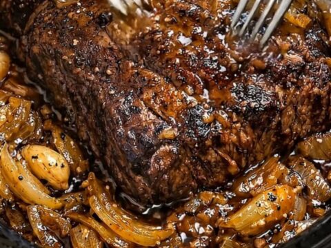 A close-up of a slow-cooked beef roast in a black slow cooker, surrounded by rich caramelized onions.