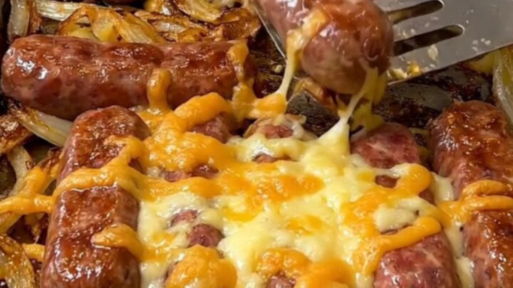 A sheet pan filled with golden-brown bratwurst, roasted baby potatoes, caramelized onion wedges, and melted cheese curds.