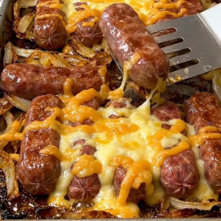 A sheet pan filled with golden-brown bratwurst, roasted baby potatoes, caramelized onion wedges, and melted cheese curds.