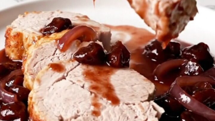A close-up of sliced pork tenderloin with dark cherry Dijon sauce and soft red onions on a white plate.