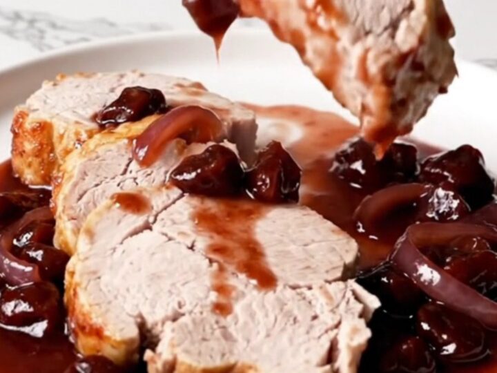 A close-up of sliced pork tenderloin with dark cherry Dijon sauce and soft red onions on a white plate.