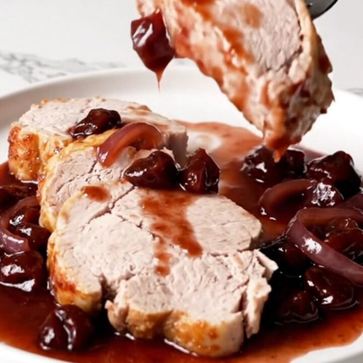 A close-up of sliced pork tenderloin with dark cherry Dijon sauce and soft red onions on a white plate.