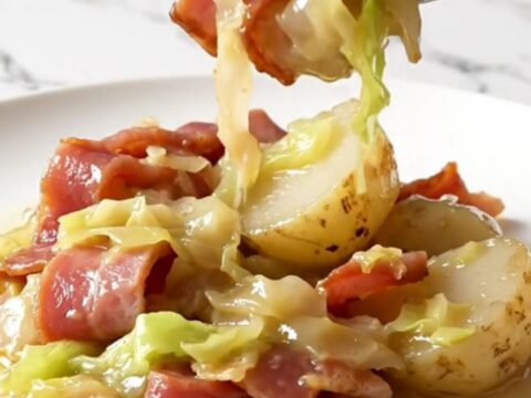 A close-up of a slow cooker filled with layers of Canadian bacon, cabbage, and baby potatoes in a buttery broth.