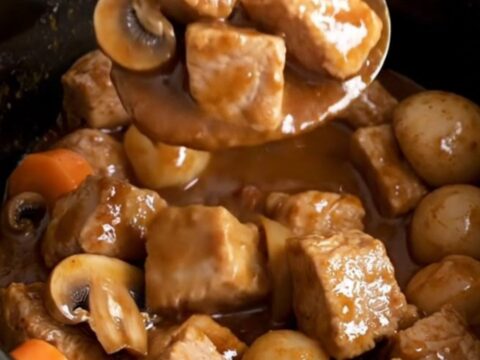 A close-up of a slow cooker filled with tender pork cubes, carrots, mushrooms, and pearl onions in a rich burgundy sauce.