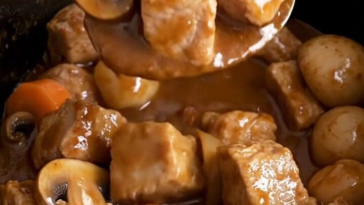 A close-up of a slow cooker filled with tender pork cubes, carrots, mushrooms, and pearl onions in a rich burgundy sauce.