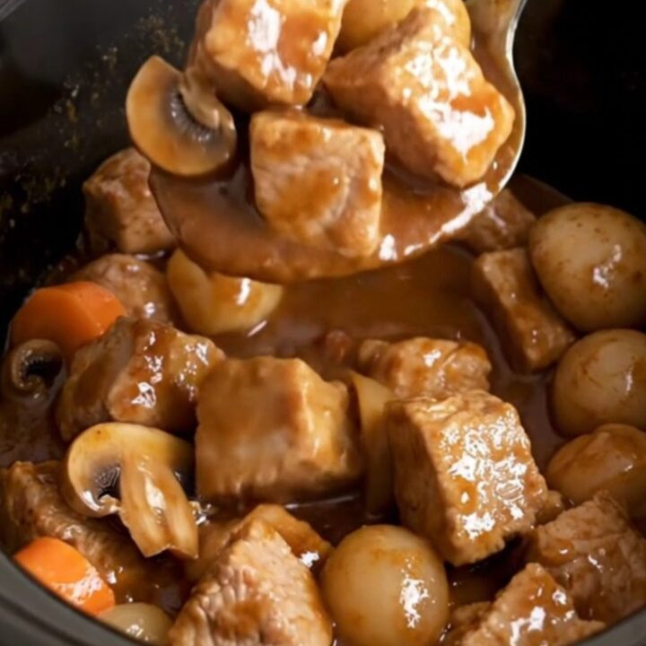 A close-up of a slow cooker filled with tender pork cubes, carrots, mushrooms, and pearl onions in a rich burgundy sauce.