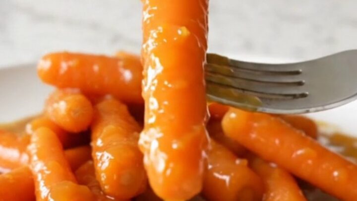 A top-down view of bright orange baby carrots inside a black slow cooker insert with a light dusting of zest and spices.