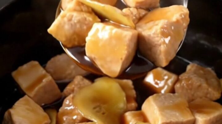 A close-up of tender, glazed pork bites in a slow cooker, featuring a rich dark brown sauce and golden ginger coins.