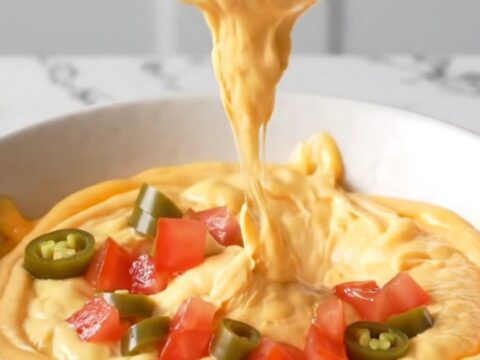 A close-up view of creamy orange queso dip topped with browned beef crumbles and diced tomatoes in a white bowl.