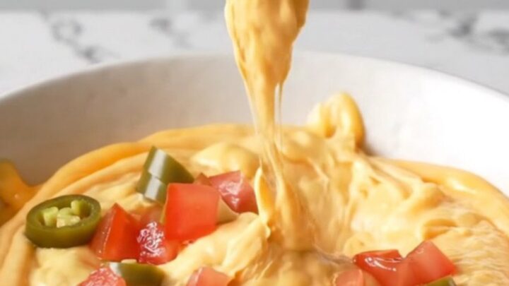 A close-up view of creamy orange queso dip topped with browned beef crumbles and diced tomatoes in a white bowl.