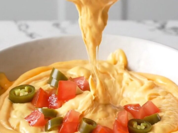 A close-up view of creamy orange queso dip topped with browned beef crumbles and diced tomatoes in a white bowl.