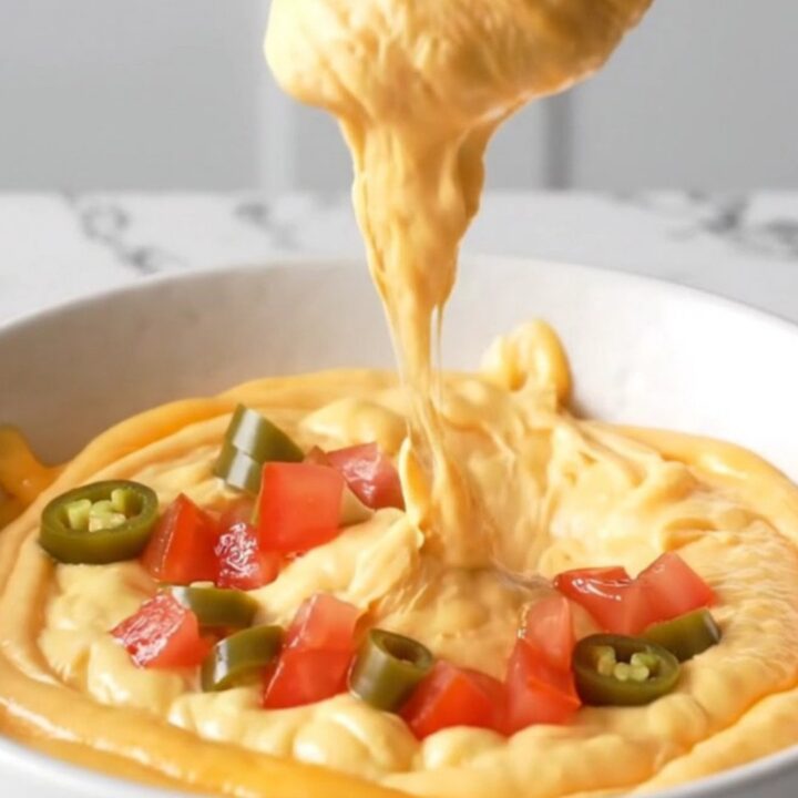 A close-up view of creamy orange queso dip topped with browned beef crumbles and diced tomatoes in a white bowl.