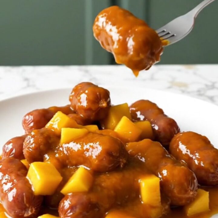 A close-up of mini smoked sausages coated in a glossy orange mango habanero glaze on a white plate.