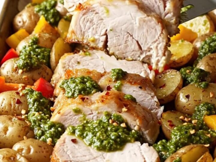 A sheet pan filled with roasted pork tenderloin slices, baby potatoes, and red bell peppers topped with green chimichurri.