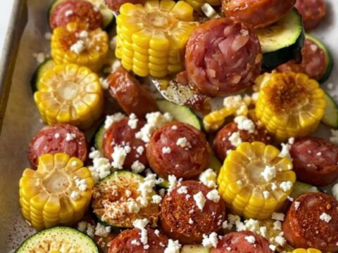 A sheet pan filled with golden-brown smoked sausage, bright green zucchini slices, and vibrant yellow corn wheels.