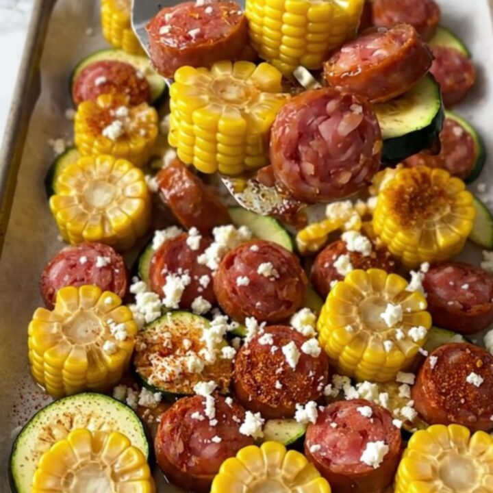 A sheet pan filled with golden-brown smoked sausage, bright green zucchini slices, and vibrant yellow corn wheels.