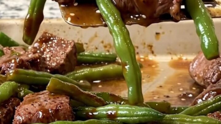 A sheet pan filled with golden-brown beef cubes, blistered green beans, and charred onion wedges, finished with sesame seeds.