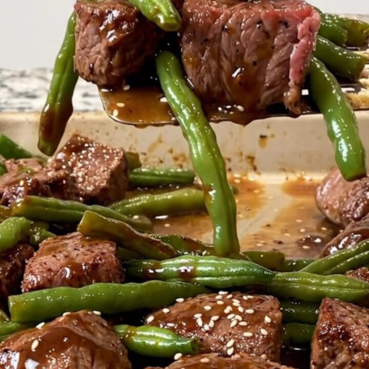 A sheet pan filled with golden-brown beef cubes, blistered green beans, and charred onion wedges, finished with sesame seeds.