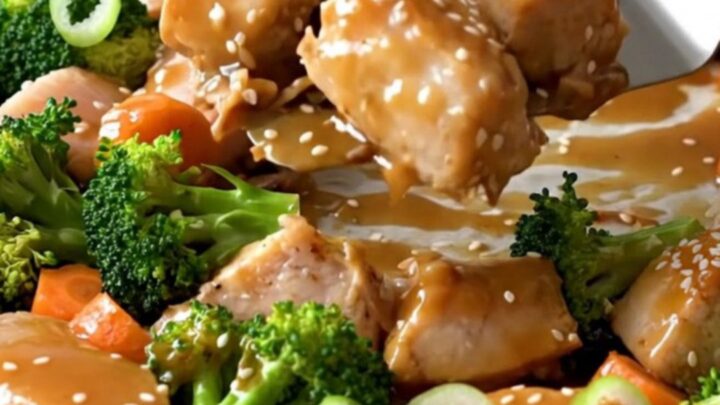 A sheet pan filled with golden-tan roasted chicken chunks, bright green broccoli florets, and orange carrot coins.
