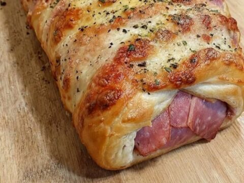 A golden-brown baked ham and cheese stromboli resting on a wooden cutting board with a slice showing melted cheese.