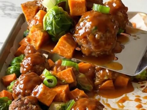 A sheet pan filled with golden turkey meatballs, roasted sweet potatoes, and Brussels sprouts drizzled in a rich red sauce.