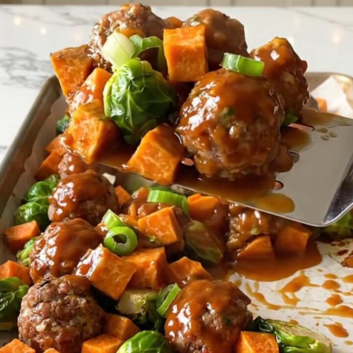 A sheet pan filled with golden turkey meatballs, roasted sweet potatoes, and Brussels sprouts drizzled in a rich red sauce.