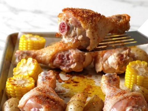 A sheet pan filled with roasted chicken drumsticks, corn on the cob, and halved baby potatoes, all golden and seasoned.