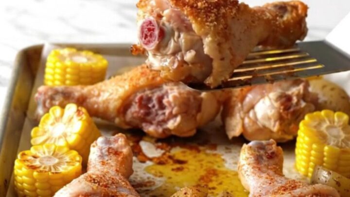 A sheet pan filled with roasted chicken drumsticks, corn on the cob, and halved baby potatoes, all golden and seasoned.