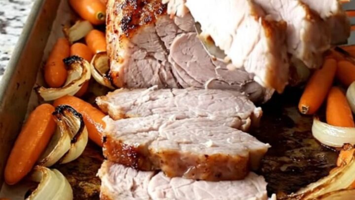 A sheet pan filled with roasted pork tenderloin, baby carrots, and red onion wedges with a dark, savory crust.