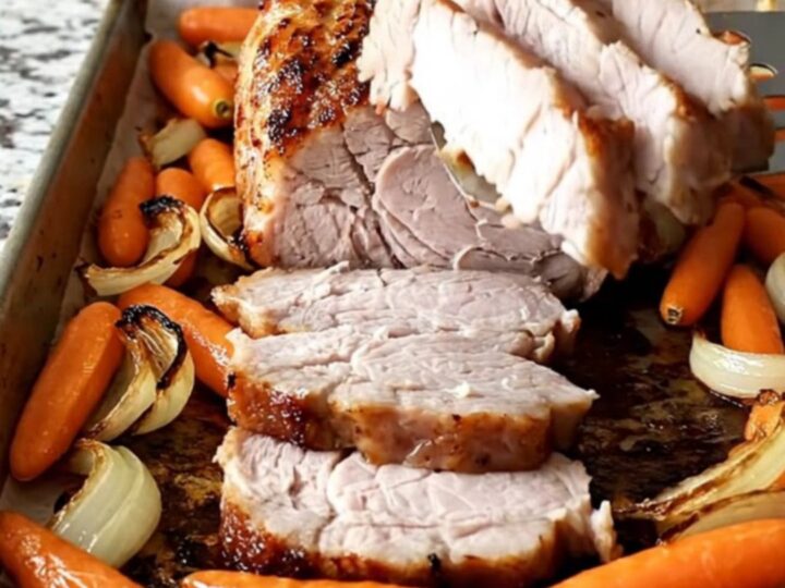 A sheet pan filled with roasted pork tenderloin, baby carrots, and red onion wedges with a dark, savory crust.