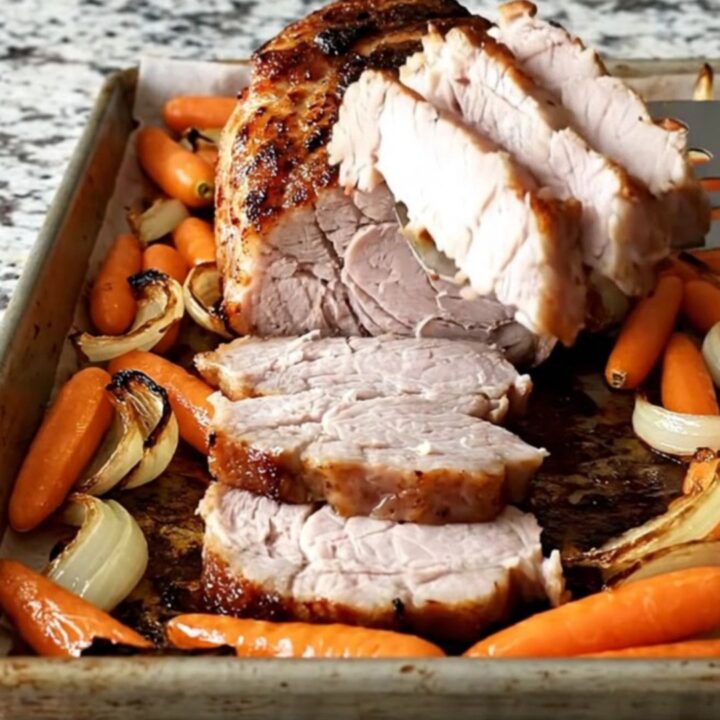 A sheet pan filled with roasted pork tenderloin, baby carrots, and red onion wedges with a dark, savory crust.