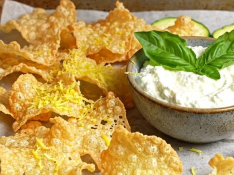 A close-up of crispy, golden-brown parmesan zucchini chips stacked next to a bowl of creamy whipped basil ricotta.