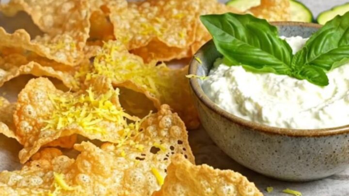 A close-up of crispy, golden-brown parmesan zucchini chips stacked next to a bowl of creamy whipped basil ricotta.