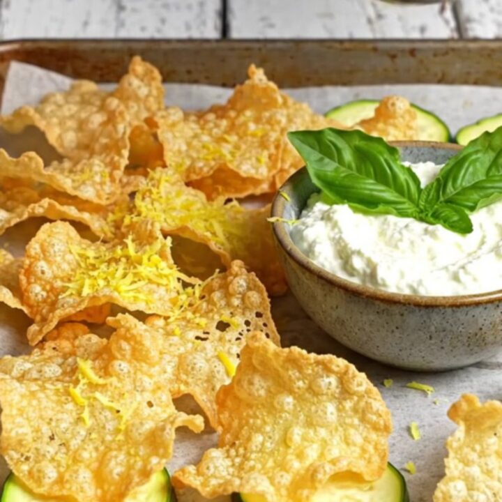 A close-up of crispy, golden-brown parmesan zucchini chips stacked next to a bowl of creamy whipped basil ricotta.