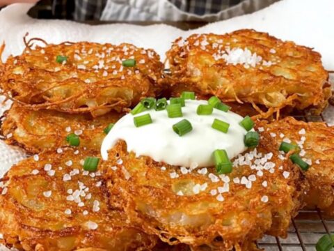 A stack of golden-brown, lacy potato latkes topped with cool sour cream and fresh green chives.