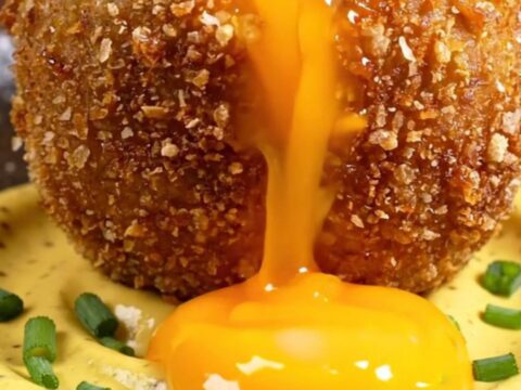A golden-fried Scotch-style potato bomb sliced in half, revealing a perfectly jammy, molten orange egg yolk flowing out.