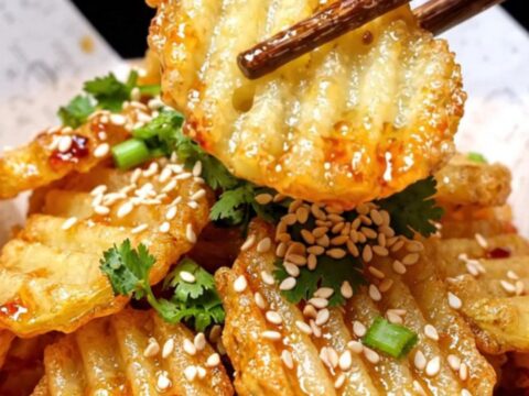 A close-up of crispy, golden-brown crinkle-cut potatoes glistening with chili oil and sprinkled with sesame seeds.