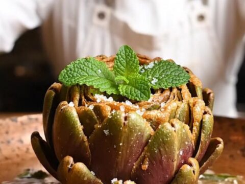 A golden, crispy Roman fried artichoke served on a rustic wooden table, with petals spread like a blooming flower.