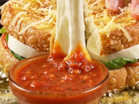 A golden-fried potato-crusted mozzarella sandwich being pulled apart with a long stretch of molten cheese.